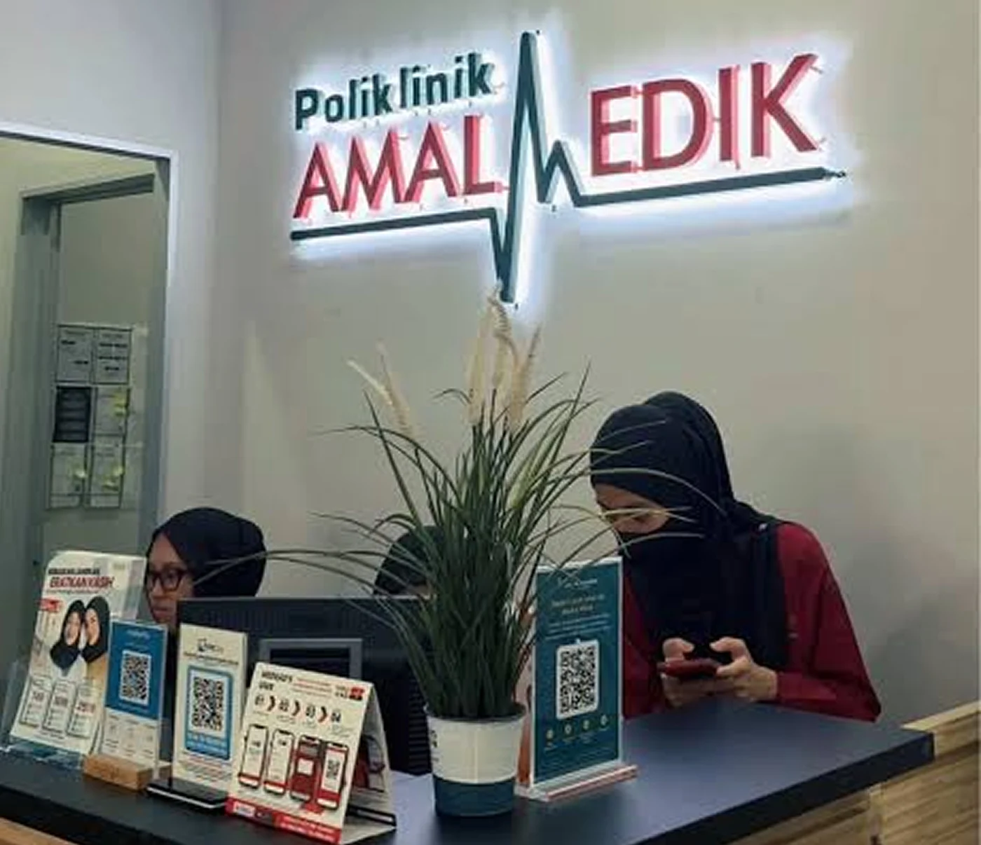 The reception counter of a Poliklinik Amalmedik clinic, the clinic's illuminated logo on the wall above it and two staff members at the desk.