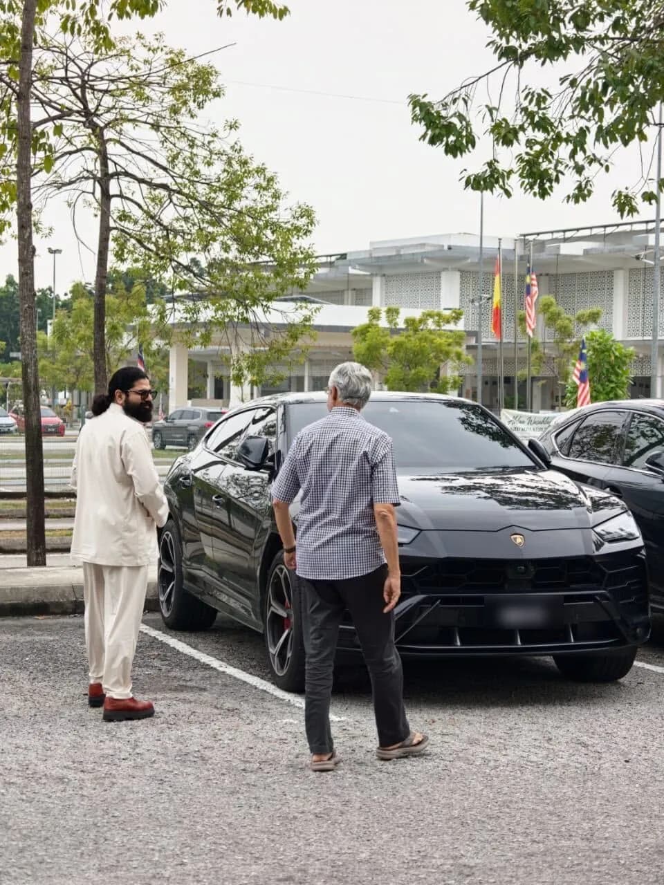 Two men talking beside a black Lamborghini Urus in a car park, with a building flying the Selangor and Malaysian flags behind them. The number plate is blurred.