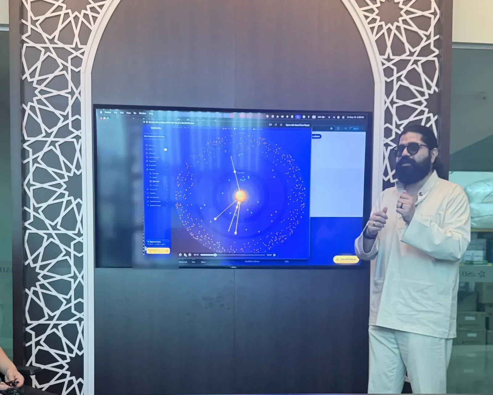 A member of the Watson team standing beside a wall screen showing Watson, explaining it to the room.