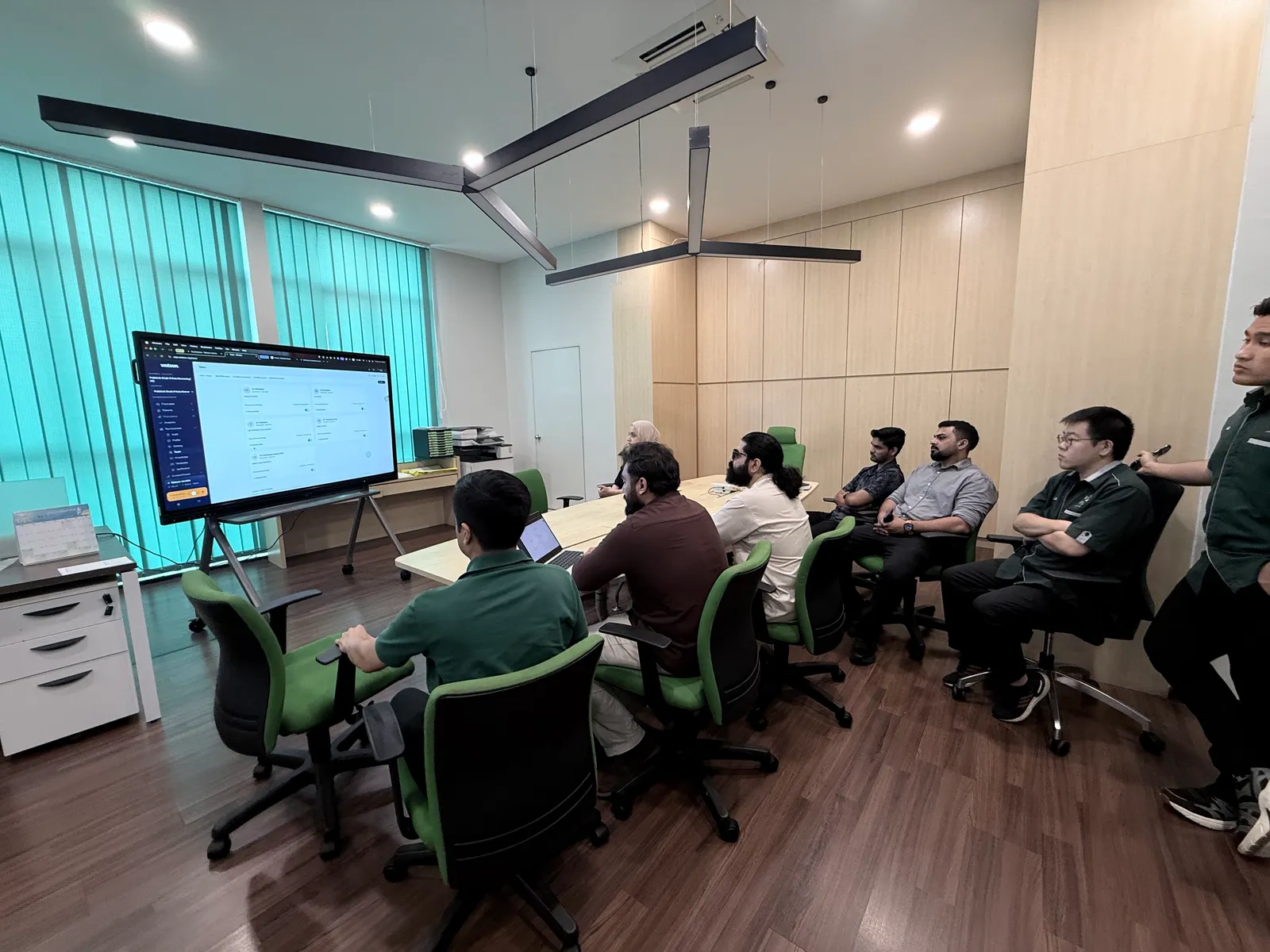 The Watson dashboard on a large screen at the front of a meeting room, with the Watson and Poliklinik Shaik teams seated at the table.