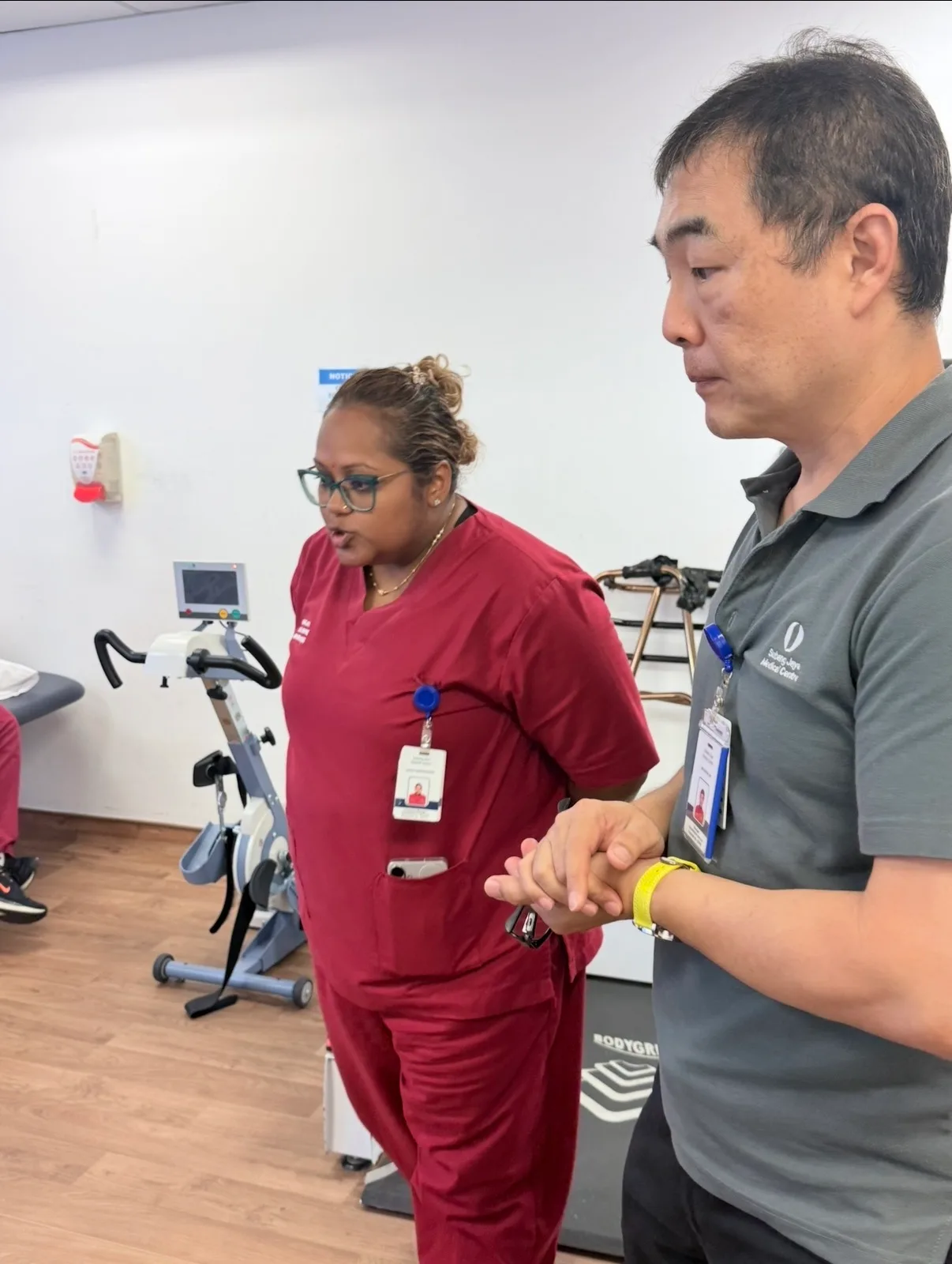 Two members of the Rehabilitative Services team standing in the rehabilitation gym at Subang Jaya Medical Centre, an exercise bike and walking frame behind them.